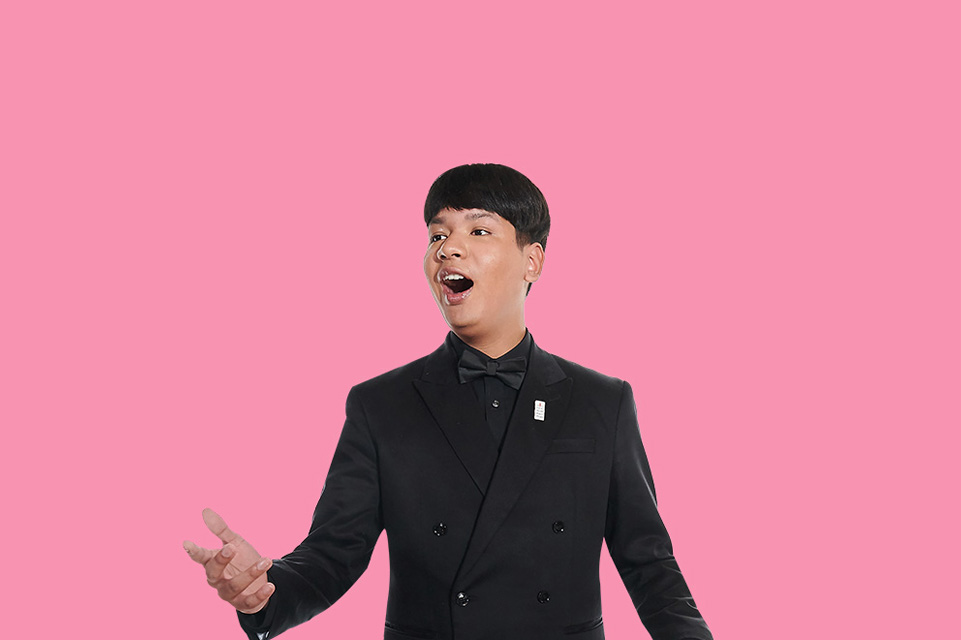 A man wearing a black double-sided blazer, with short hair, singing, with a light pink background.